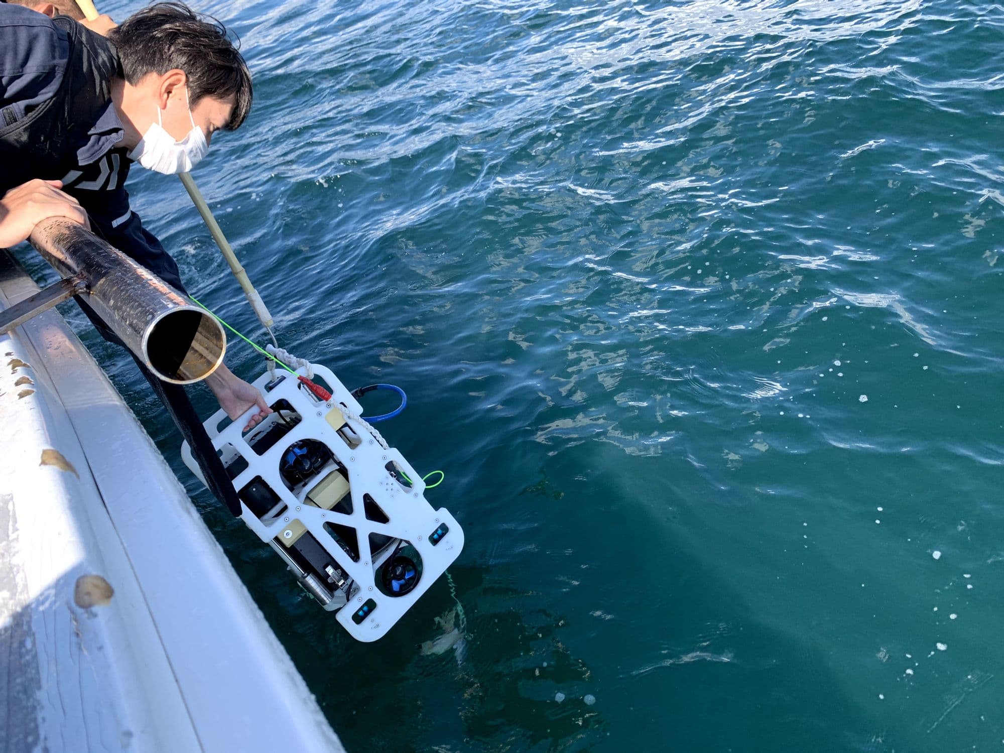 一人は水中ドローンを手で持ち、もう一人は先端にかぎ針のついた棒を使って、着水の衝撃を与えないように静かに投入します。